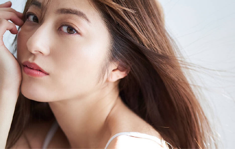 風になびくブラウンのロングヘアが美しい女性のポートレート。透明感のある肌とナチュラルな表情で、柔らかい自然光が差している。