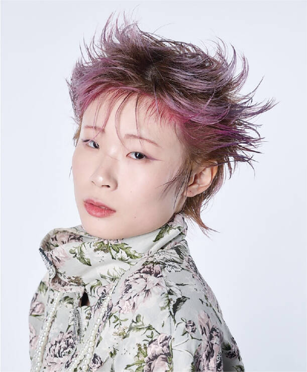 鮮やかなピンクとブラウンを組み合わせた立体感あるショートヘアが印象的で、花柄のハイネック衣装とともに独創的な美を表現している。
